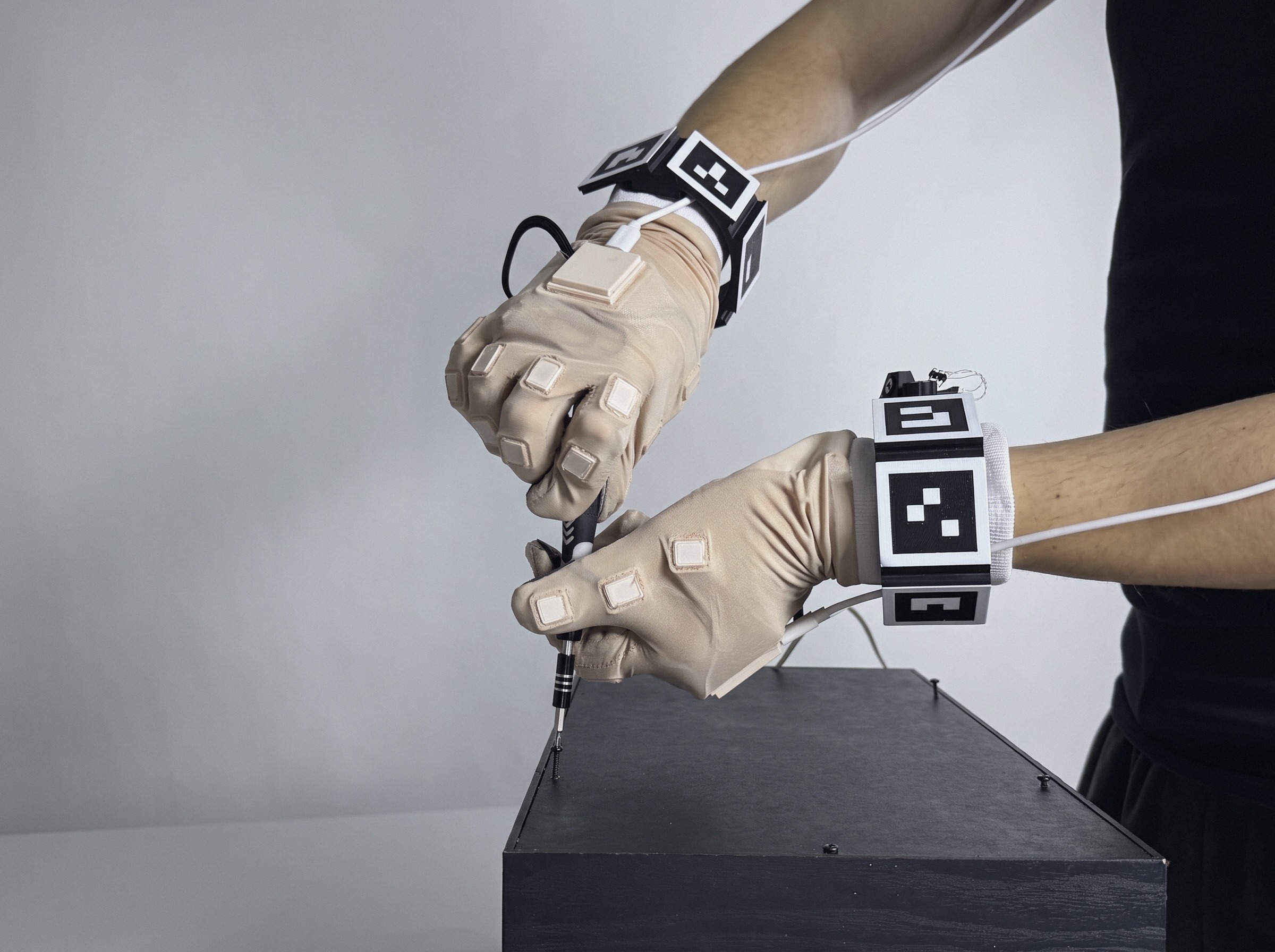 Two hands wearing the April Glove with wrist tracking modules drive a screw into a black housing with a precision screwdriver.